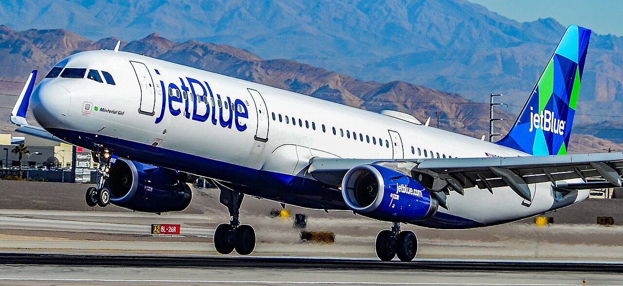 A JetBlue passenger airplane taking off from a runway with mountains in the background.