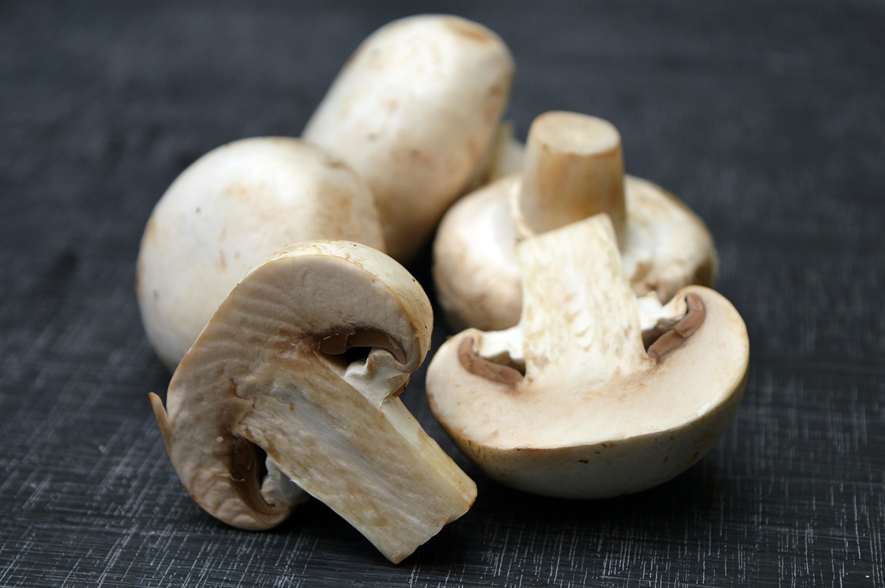 A close-up of several white button mushrooms, some whole and some sliced, on a dark surface.