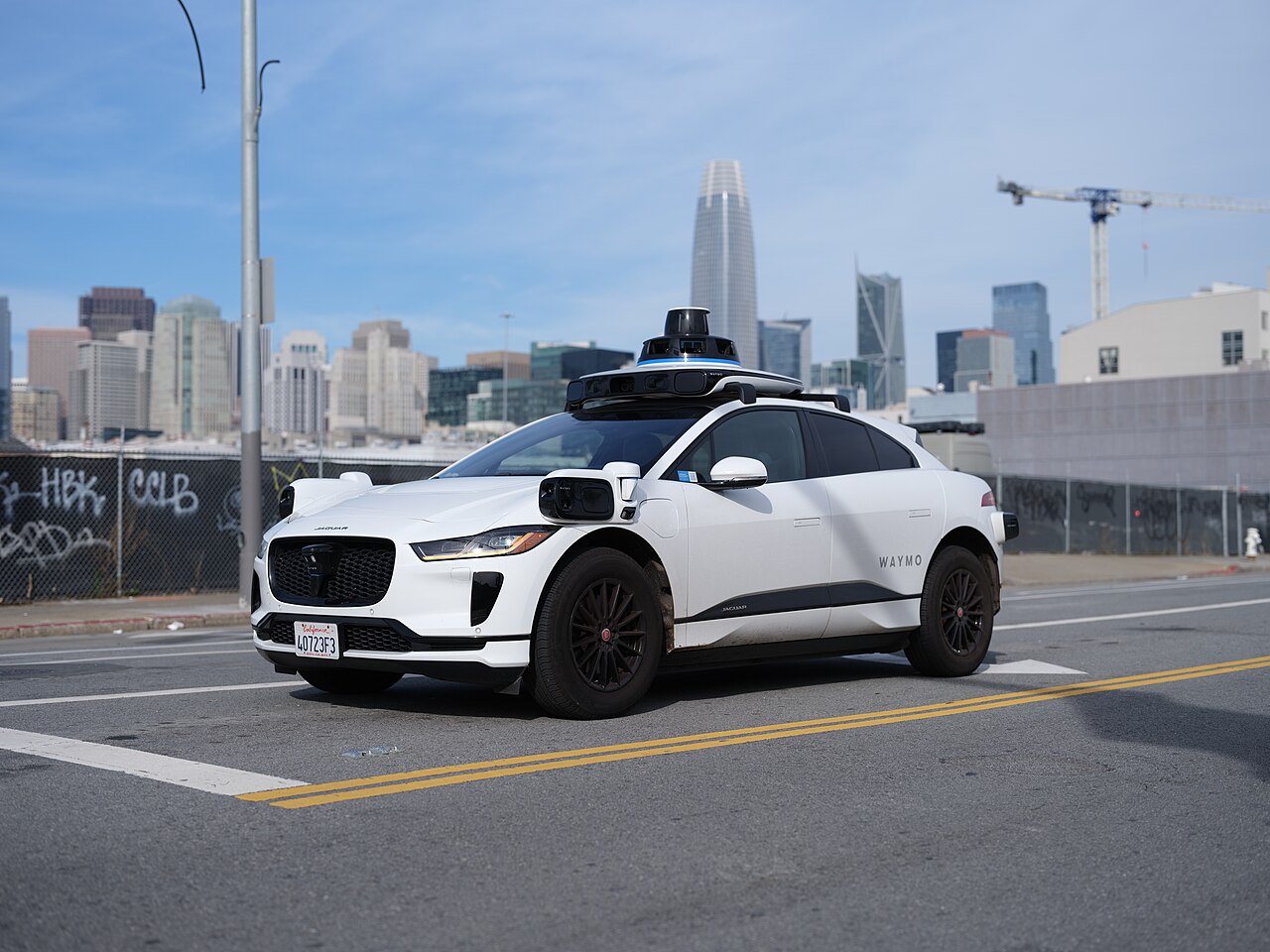 A white Waymo self-driving Jaguar I-PACE vehicle driving on a city street with a skyline in the background.