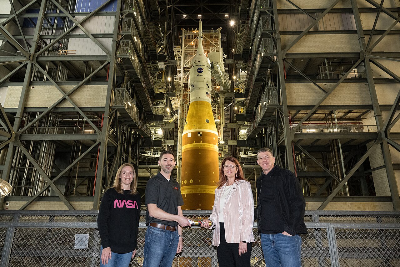 Four people standing in front of a large NASA rocket inside a hangar.