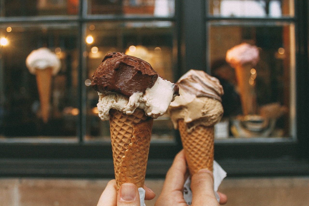 Two hands holding ice cream cones with different flavors in front of a shop window.