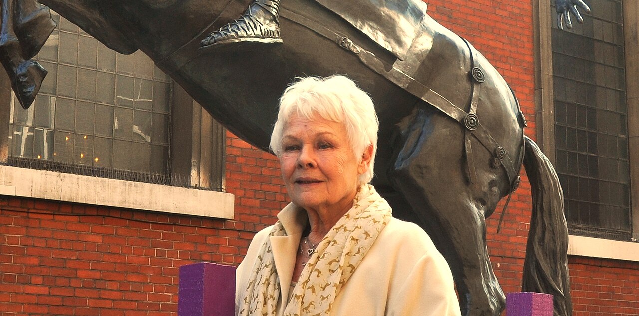 Dame Judi Dench standing in front of a bronze horse statue in a medium close-up shot.