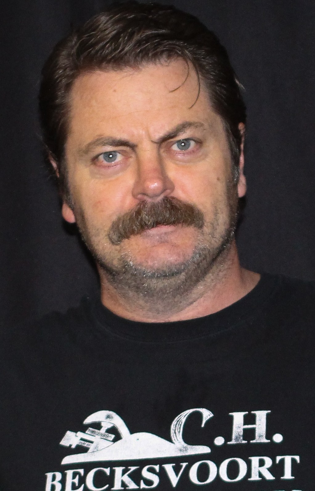 Close-up portrait of Nick Offerman with a mustache, wearing a dark t-shirt with text.