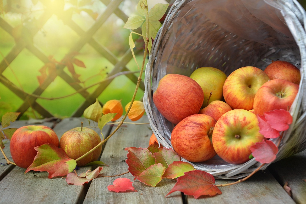 A basket tipped over on a wooden surface, spilling fresh apples surrounded by autumn leaves.