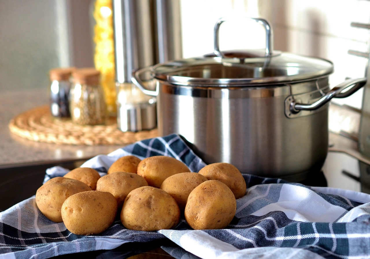 A pile of raw potatoes on a checkered cloth next to a stainless steel cooking pot in a kitchen.