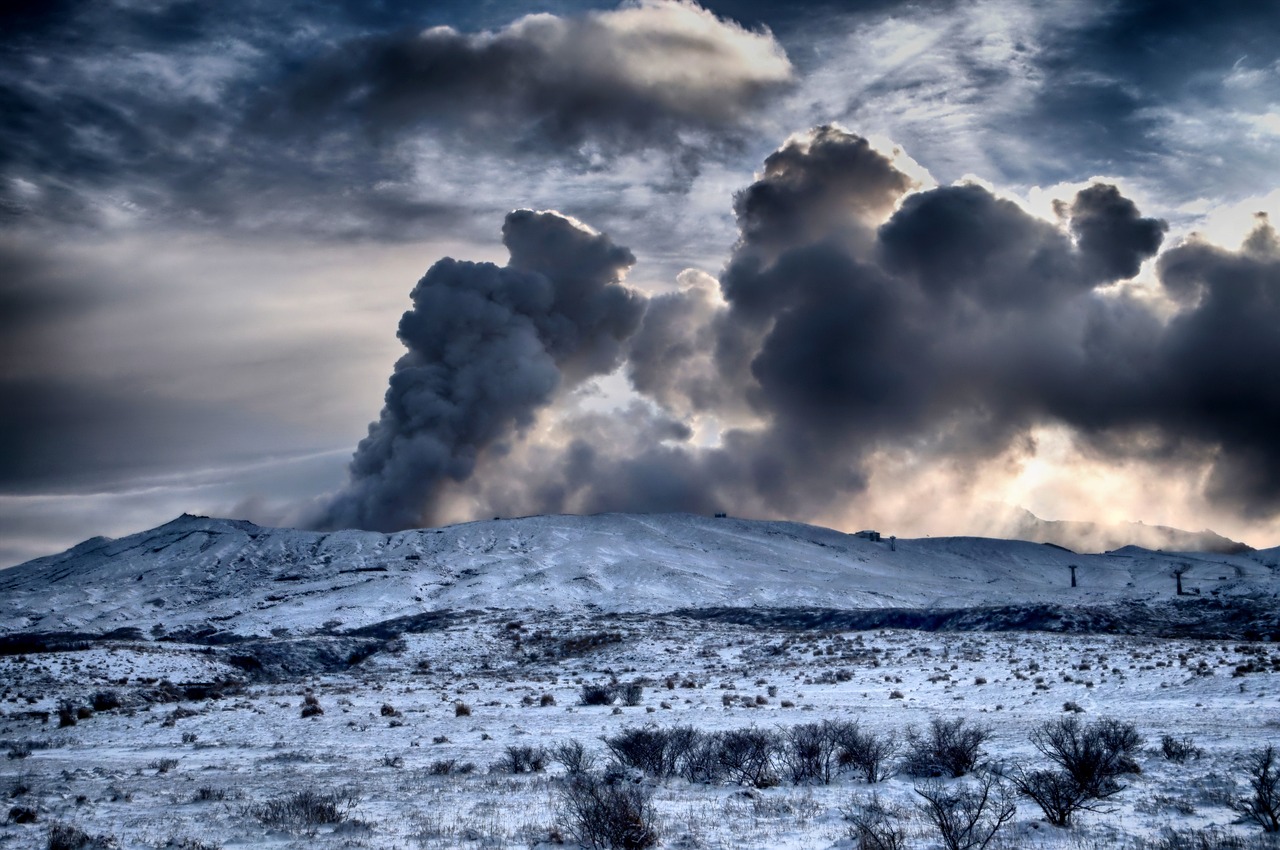 A large volcano erupting, sending a huge plume of dark smoke and ash into the cloudy sky above a snow-covered landscape.