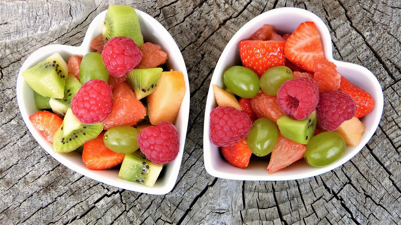 Two white heart-shaped bowls filled with mixed fresh fruit salad, including raspberries, strawberries, kiwi, grapes, and melon, on a rustic wooden surface.